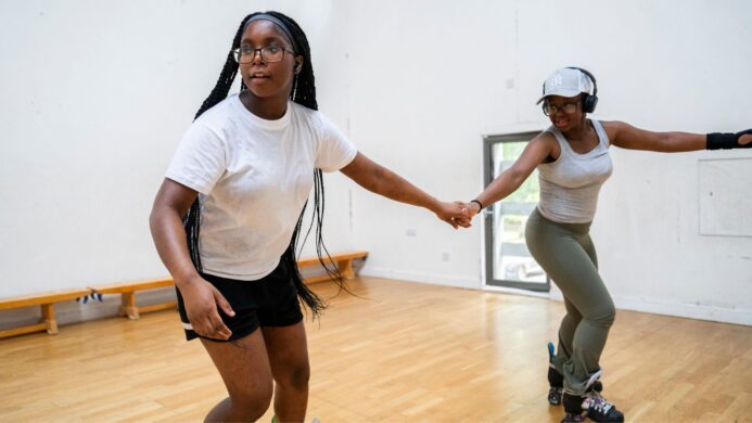 Two young women holding hands and roller skating in a sports hall